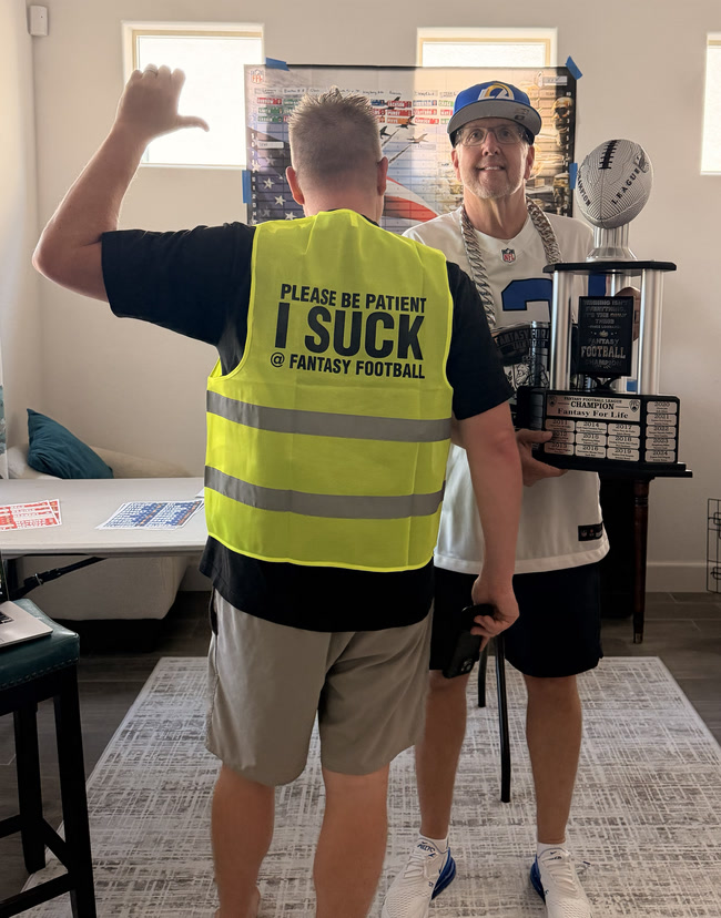 The Toilet Bowl winner pointing at the back of the new vest, which reads Please be patient, I suck at fantasy football, with the new championship trophy behind him
